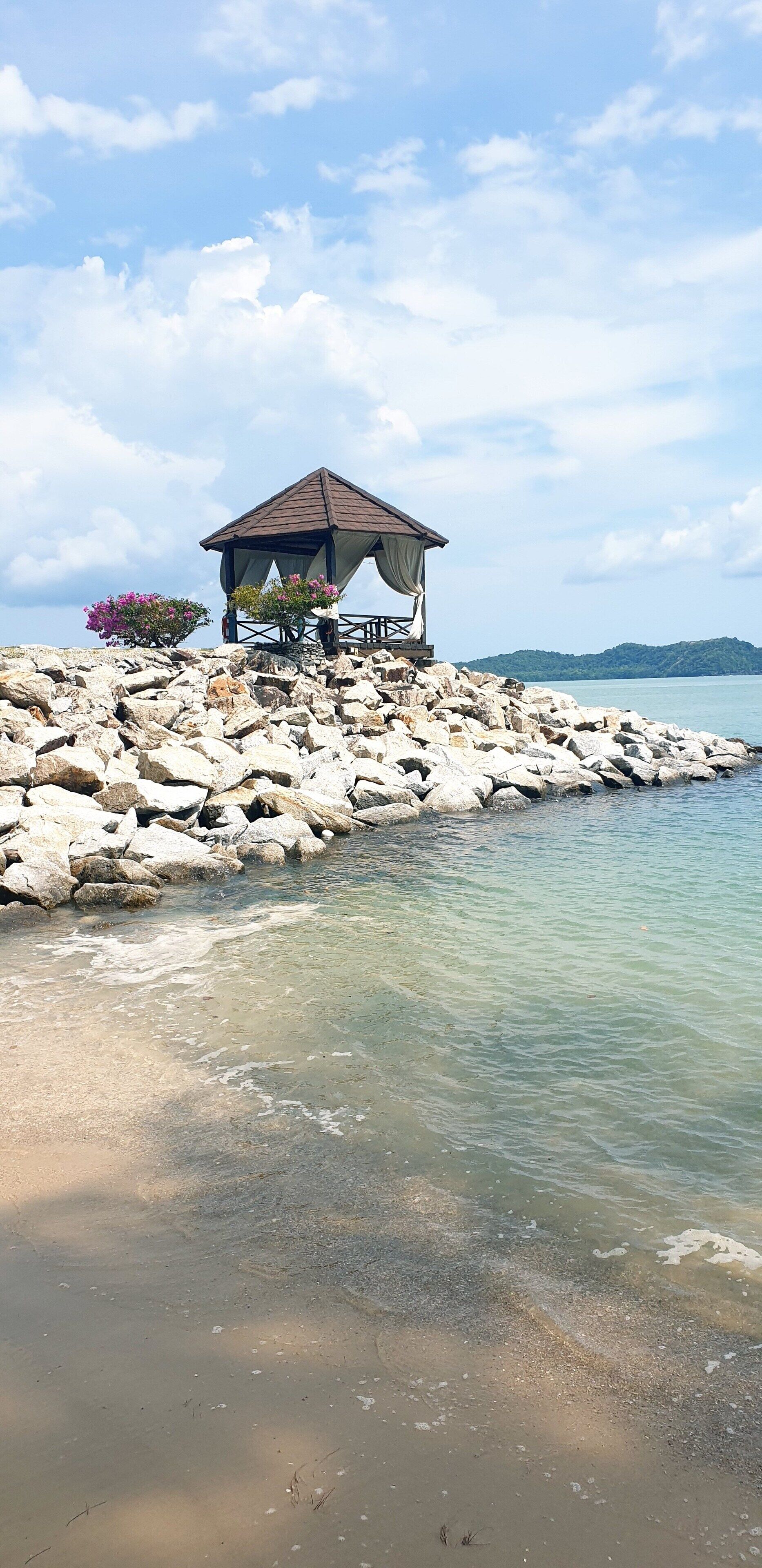 A gazebo with a roof and a table, surrounded by rocks and flowers, overlooking a calm body of water.