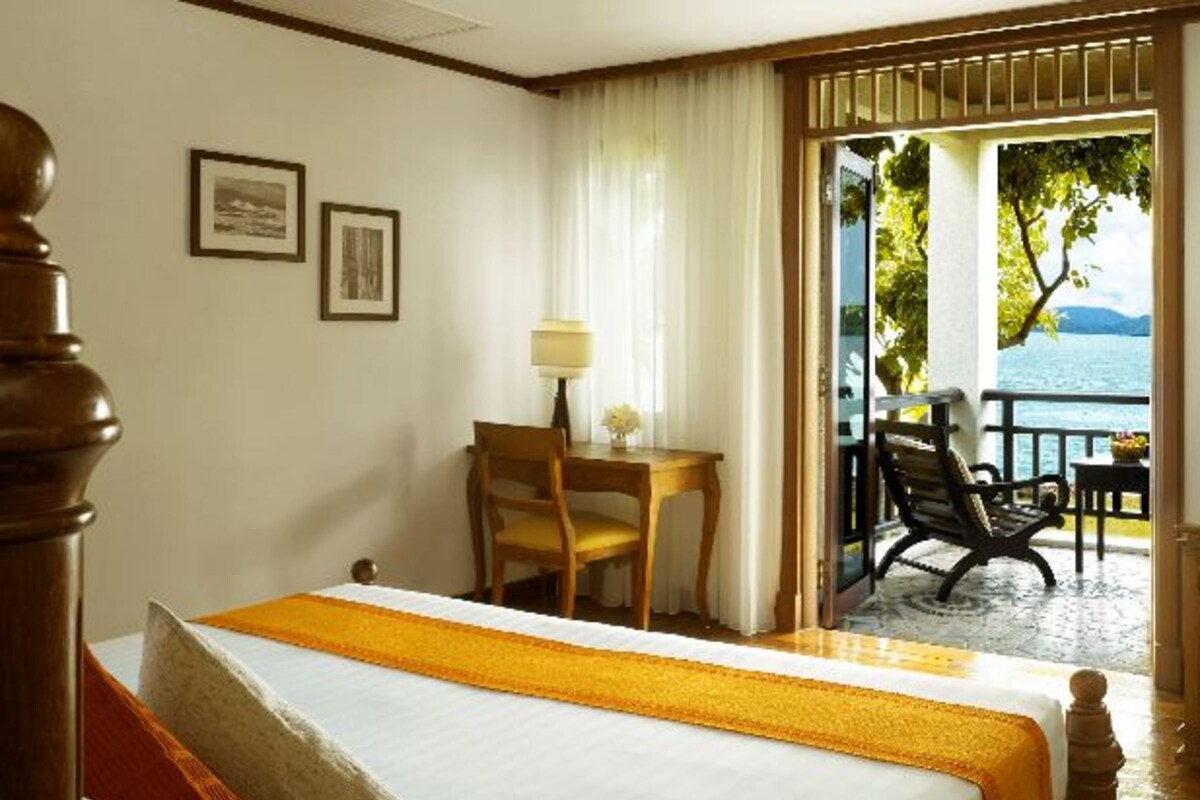A hotel room with a bed, a desk, and a chair. There is a view of the sea through the window.
