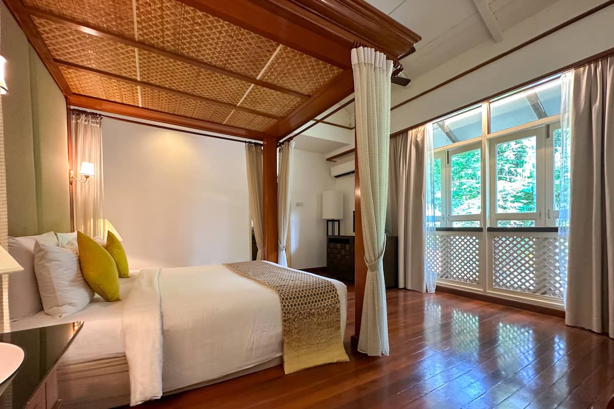 A spacious bedroom with a large bed, wooden flooring, a ceiling fan, and a view of greenery outside.