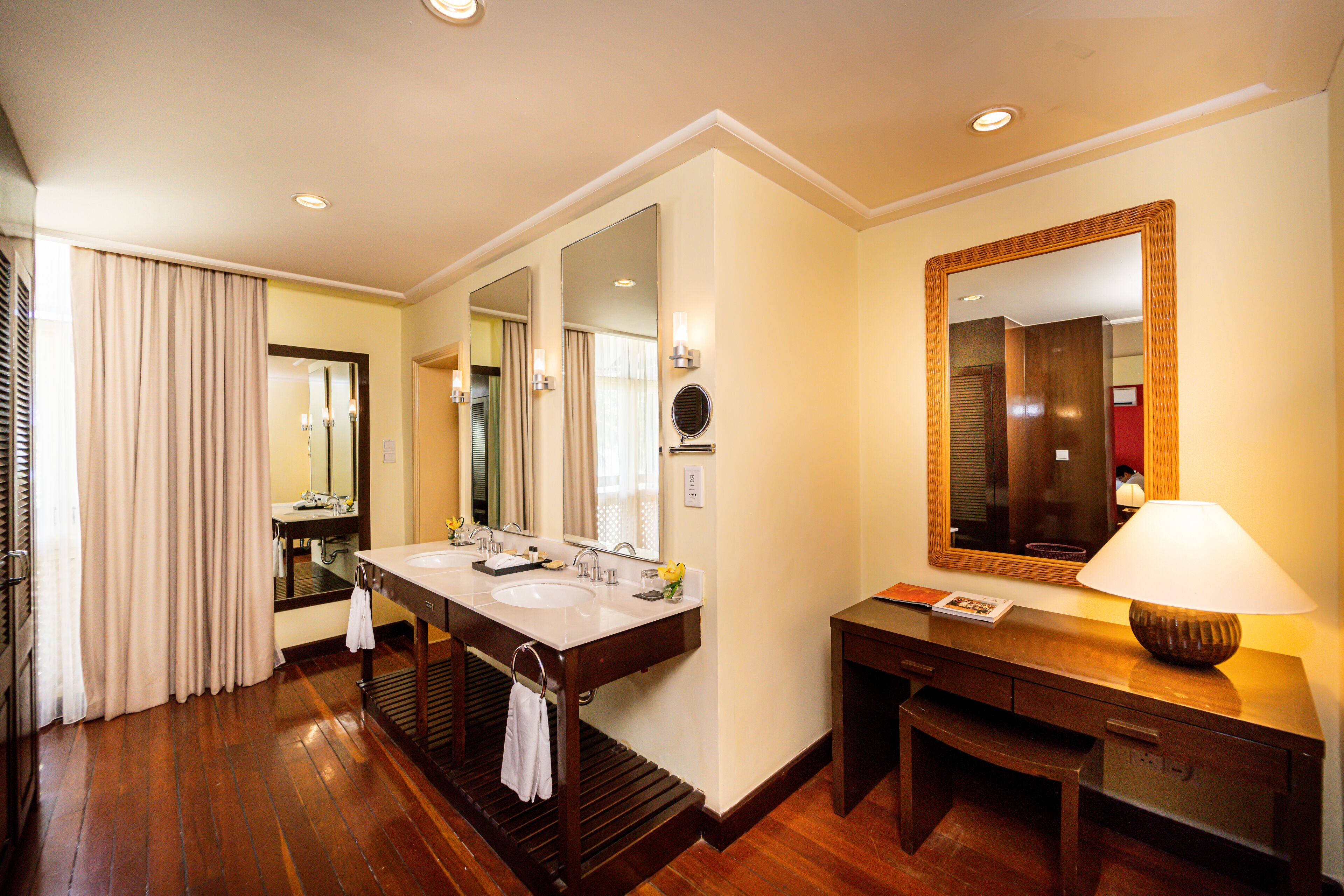 A hotel bathroom with a double sink vanity, a large mirror, and a bedside table with a lamp.