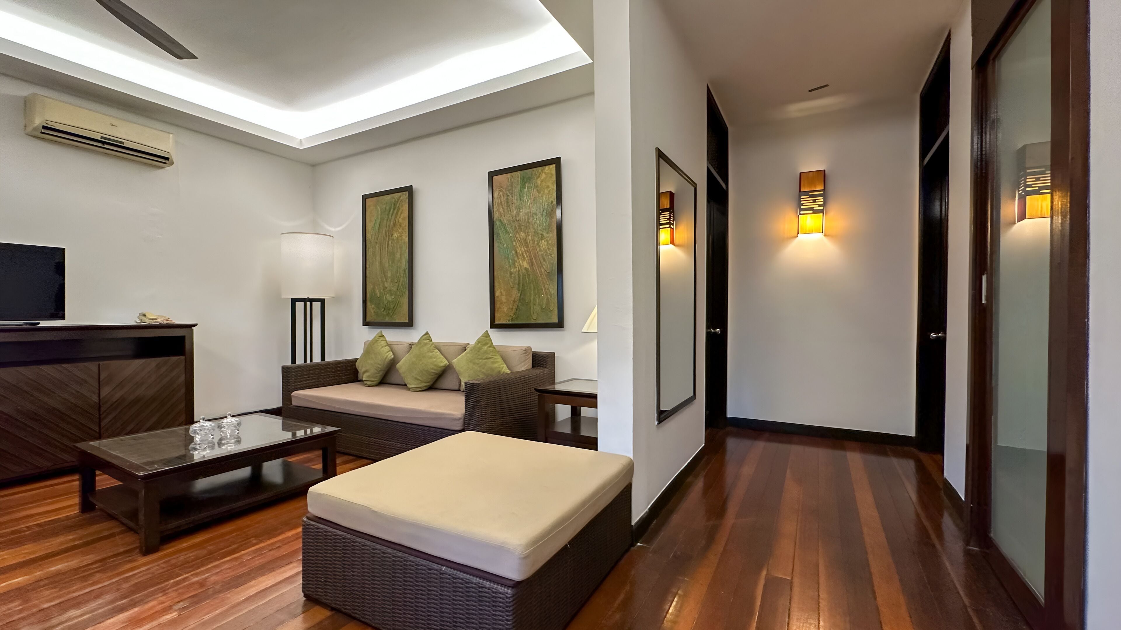 A modern hotel room with a sofa, ottoman, and a flat-screen TV on a wooden cabinet.