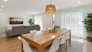 Ett modernt vardagsrum med ett matbord i trä, vita stolar, en grå soffa och ett stort fönster med tunna, genomskinliga gardiner.
