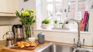 A kitchen with a stainless steel sink, a coffee maker, a kettle, a jar of coffee beans, a vase with flowers, and a few croissants on a wooden board.