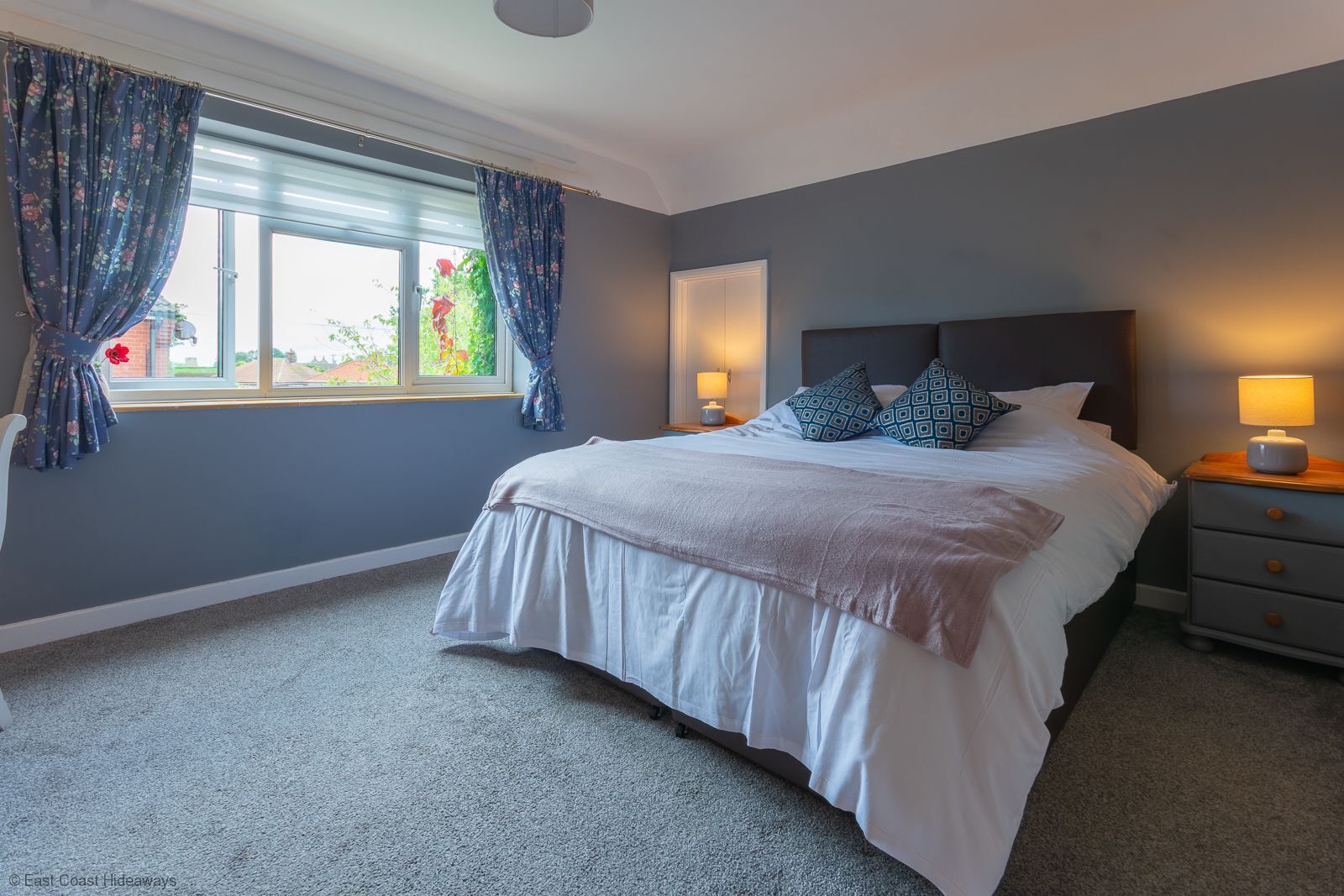 A bedroom with a large bed, two bedside tables with lamps, a window with curtains, and a grey carpet.