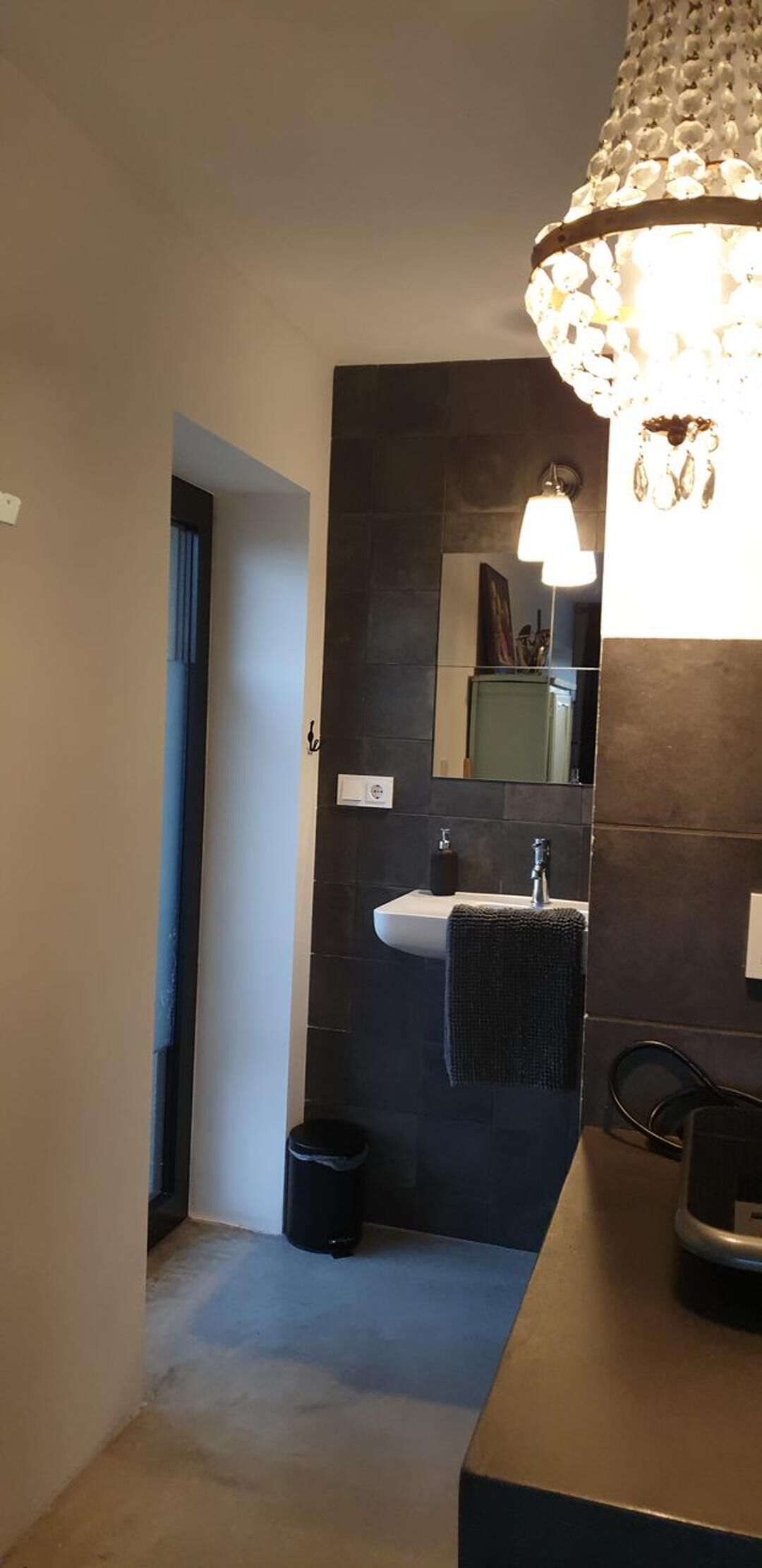 A modern bathroom with a dark tiled wall, a white sink, a mirror, and a chandelier.