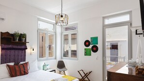 Ein modernes Hotelzimmer mit einem großen Bett, einem kleinen Schreibtisch, einem Sessel und einem Esstisch. Es gibt Fenster, eine Tür und einen Blick nach draußen.