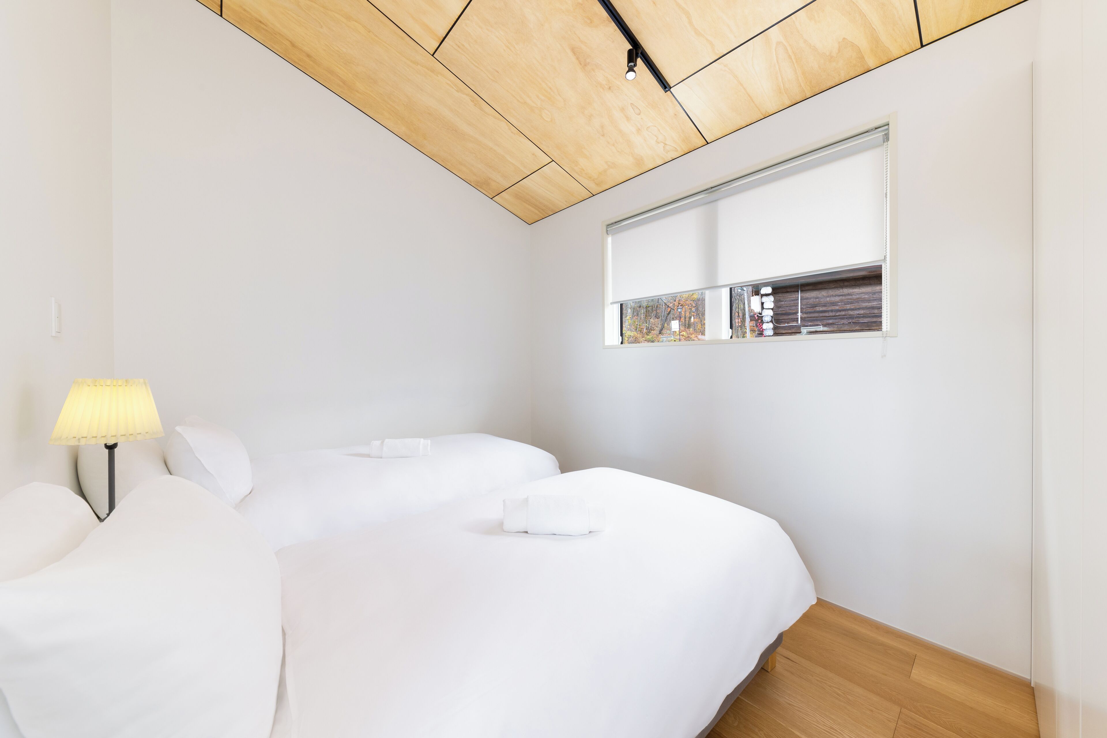A modern bedroom with a wooden ceiling, a large window with blinds, a bedside table with a lamp, and neatly made beds.