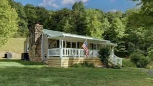 A stone house with a porch and an American flag, surrounded by a lush green landscape. - PET FRIENDLY COTTAGE 3.5 MILES FROM LAKE MOOMAW! (Hot Springs)