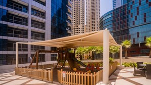 Un'area giochi all'aperto con uno scivolo e altalene, circondata da una recinzione in legno, con sullo sfondo edifici moderni.