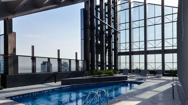 Una piscina en la azotea con agua azul cristalina, rodeada por una estructura moderna de vidrio y una barandilla de metal.