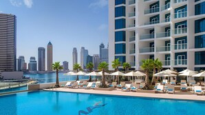 Ein Hotelpool mit Liegestühlen und Sonnenschirmen, im Hintergrund ein Stadtpanorama und ein klarer, blauer Himmel.