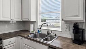 A kitchen with white cabinets, a stainless steel sink, a window with blinds, and a coffee maker. - Summer Bliss | Walk to Beach • Master Suite • Bunk Beds • Sleeps 9 (Galveston)