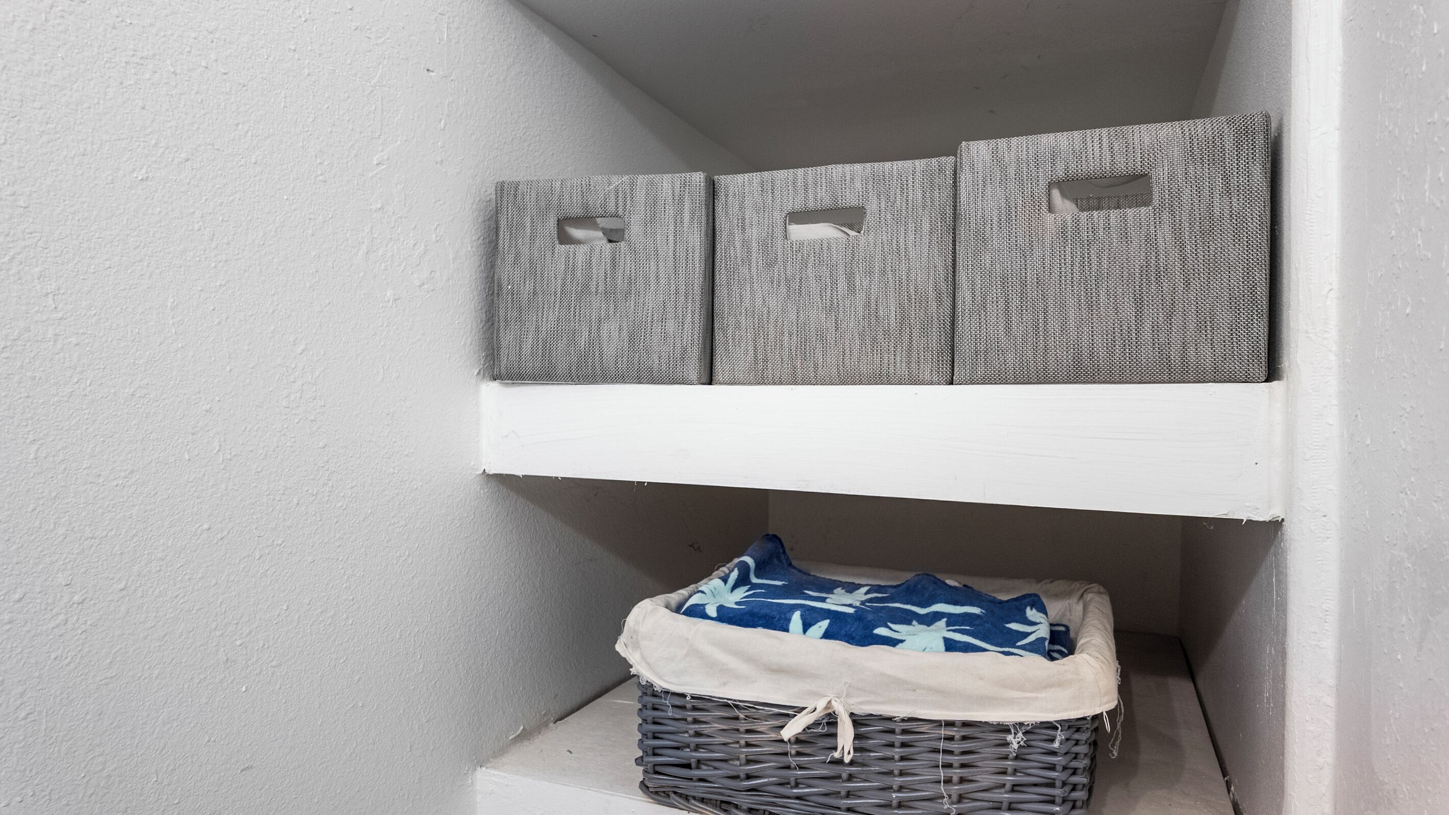 A corner of a room with a white wall, a white shelf, and a wicker basket with a blue and white blanket.