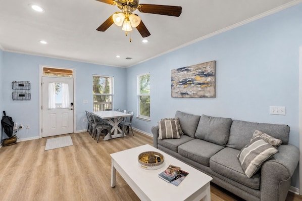 A living room with a gray sofa, a white coffee table, a dining table with chairs, a ceiling fan, and a painting on the wall.