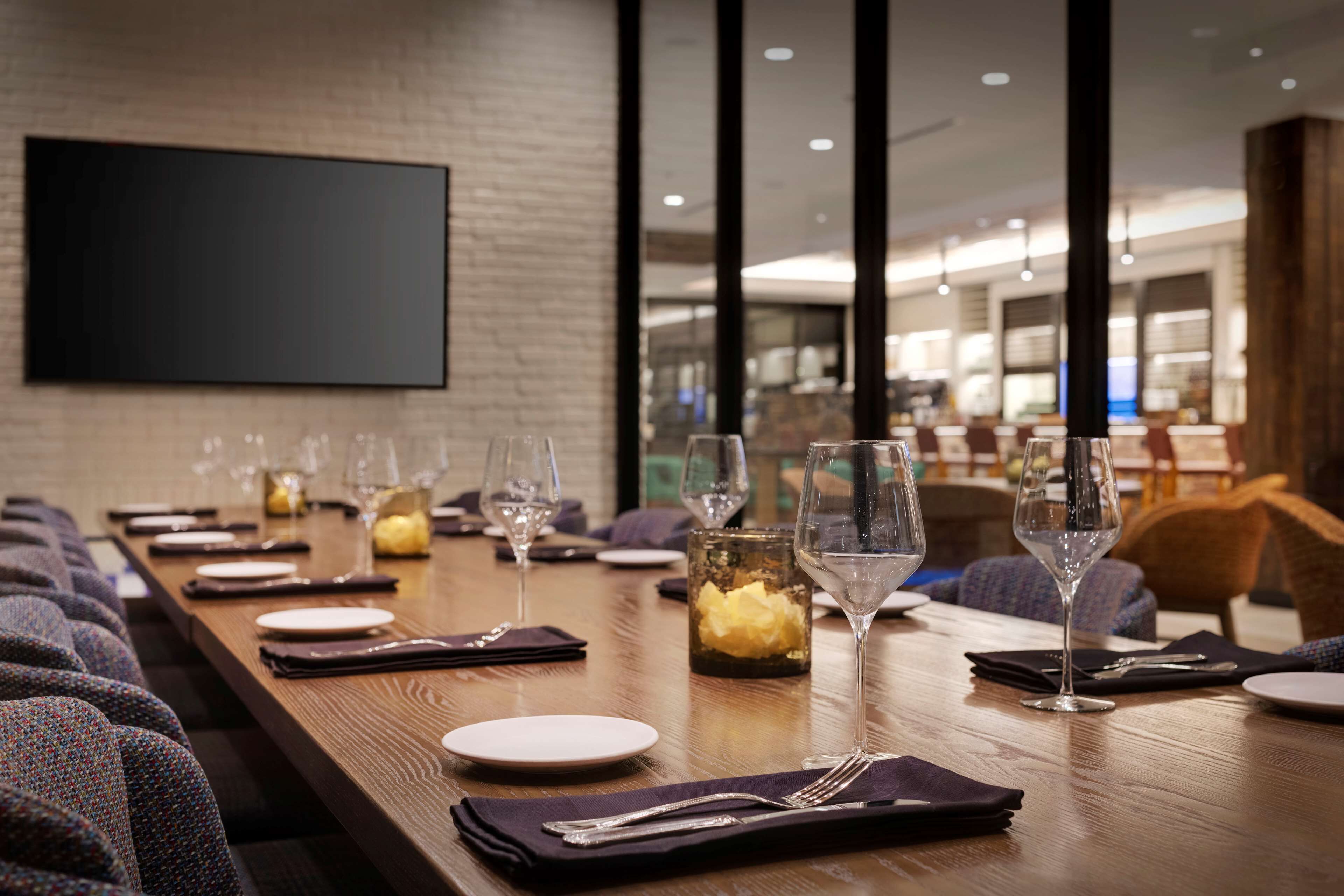 A long dining table set with plates, glasses, and cutlery, with a modern interior and a large screen on the wall.