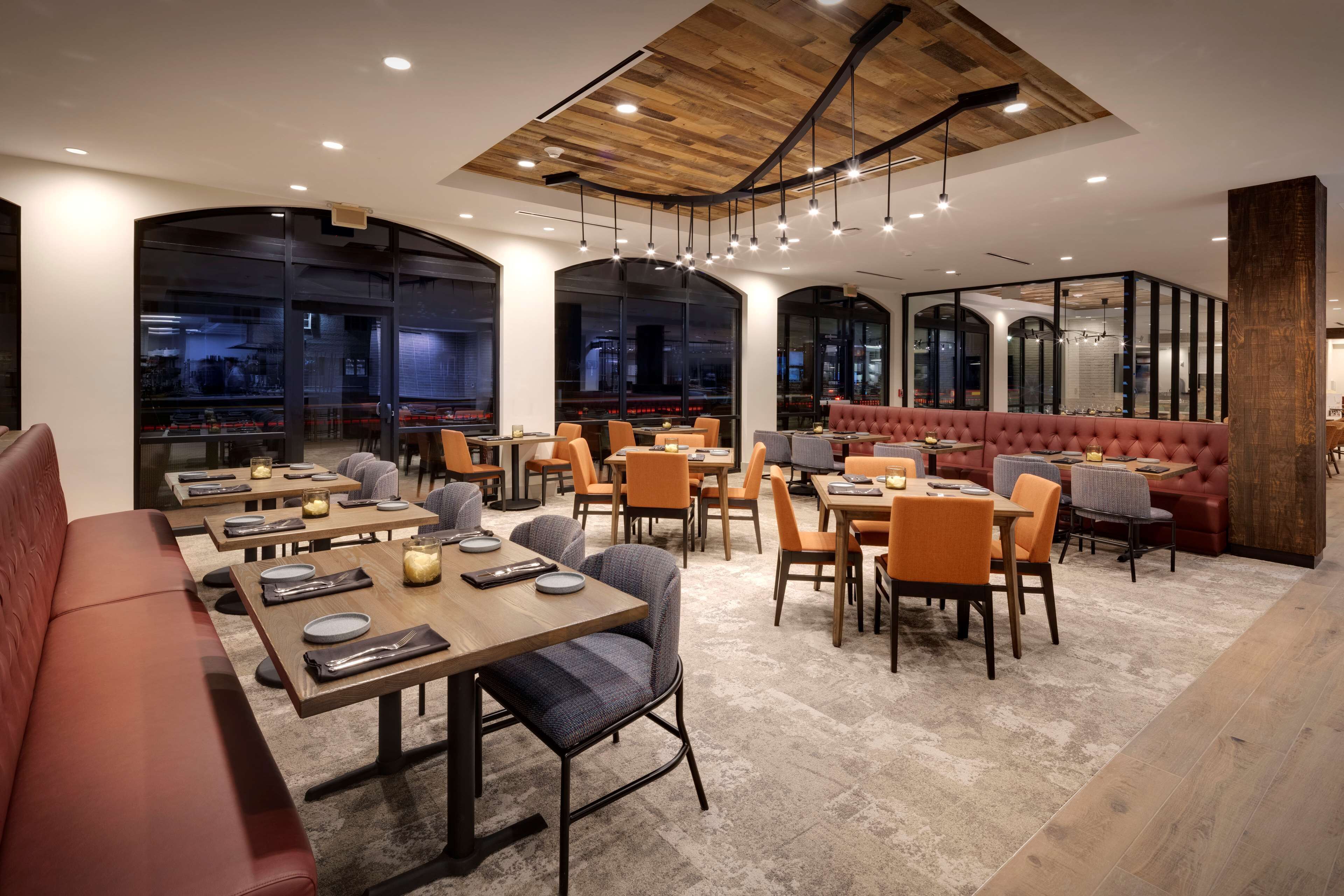 A modern restaurant with wooden tables and chairs, a red booth, and large windows offering city views.