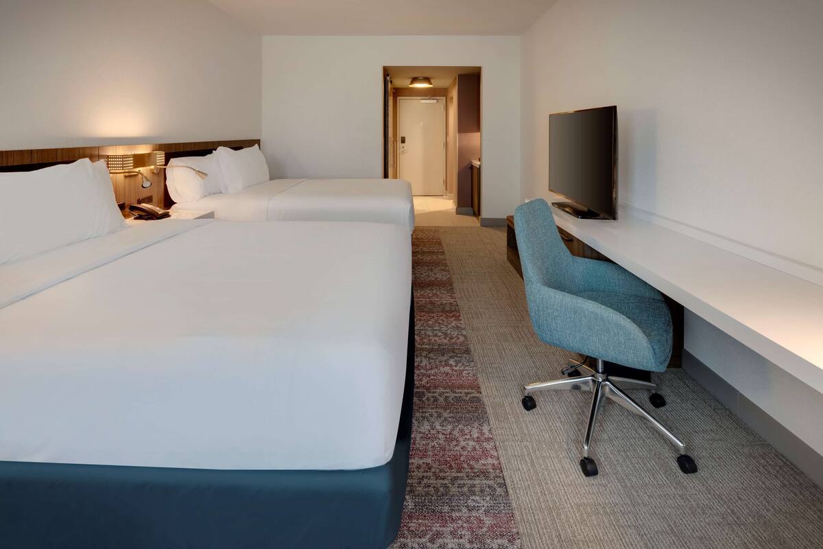 Una habitación de hotel con dos camas, un escritorio con silla, televisión y una puerta que da a otra habitación.