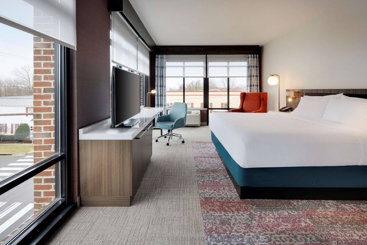 Una habitación de hotel con cama, escritorio, silla, televisor y un ventanal grande.