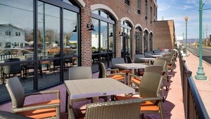 Un espace extérieur aménagé avec des chaises et des tables en osier, de grandes fenêtres et une façade en briques.