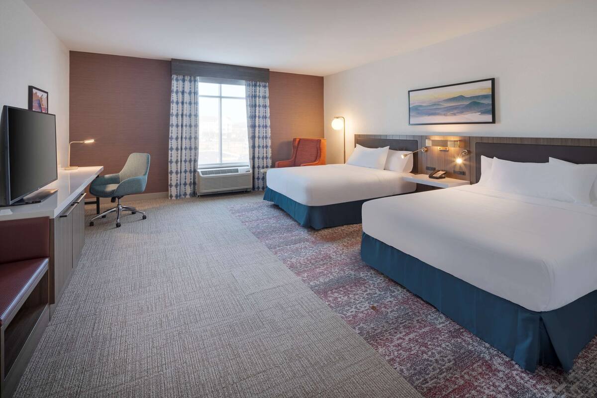 Una habitación de hotel con dos camas, un escritorio con silla, televisión y un ventanal con cortinas.