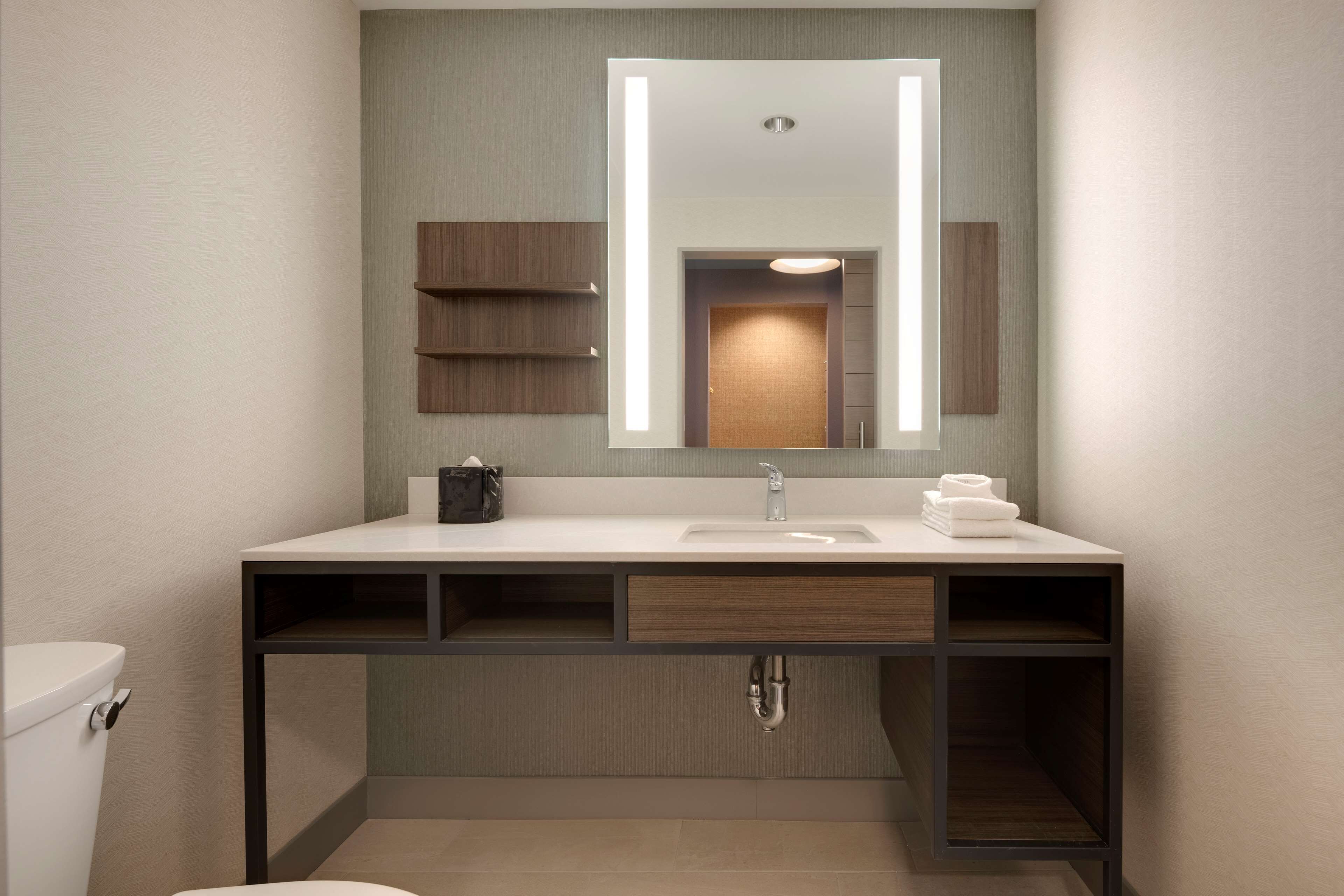 A modern bathroom with a white sink, a mirror with integrated lighting, a towel rack, and a toilet.