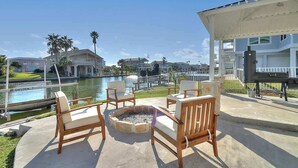 An outdoor seating area with wooden chairs and a fire pit, overlooking a body of water and buildings.