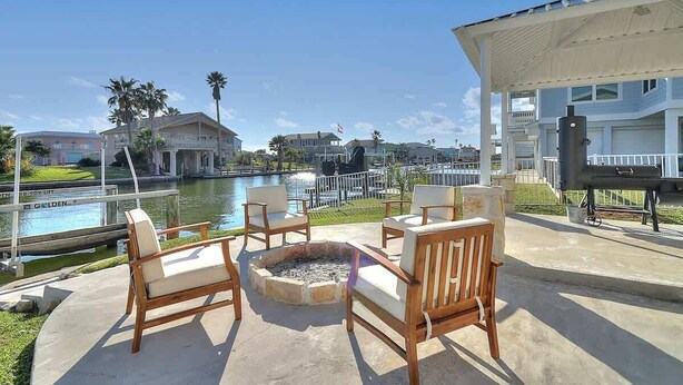An outdoor seating area with wooden chairs and a fire pit, overlooking a body of water and buildings.