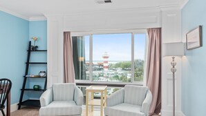 Una habitación con dos sillones a rayas, una mesita, una estantería y un amplio ventanal con vistas a un faro y barcos.