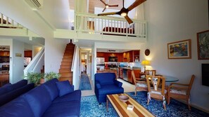 A spacious living area with a staircase leading to a balcony, a kitchen in the background, and a dining area with chairs and a table.