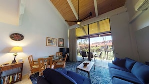 Ein geräumiges Wohnzimmer mit hohen Decken, Holzbalken, einem großen Fenster mit Blick ins Freie, einem Deckenventilator und einer Wanduhr.