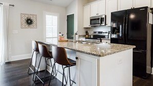 A modern kitchen with white cabinets, a black refrigerator, a microwave, and a stainless steel oven. There are three bar stools at the island and a decorative wall piece above the window. - A Home in One ⛳️ Sleeps 10 & 🎱 Table! (West End)
