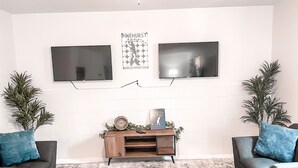 A modern living room with two black sofas, a wooden coffee table, and two large flat-screen TVs mounted on a white brick wall. - A Home in One ⛳️ Sleeps 10 & 🎱 Table! (West End)
