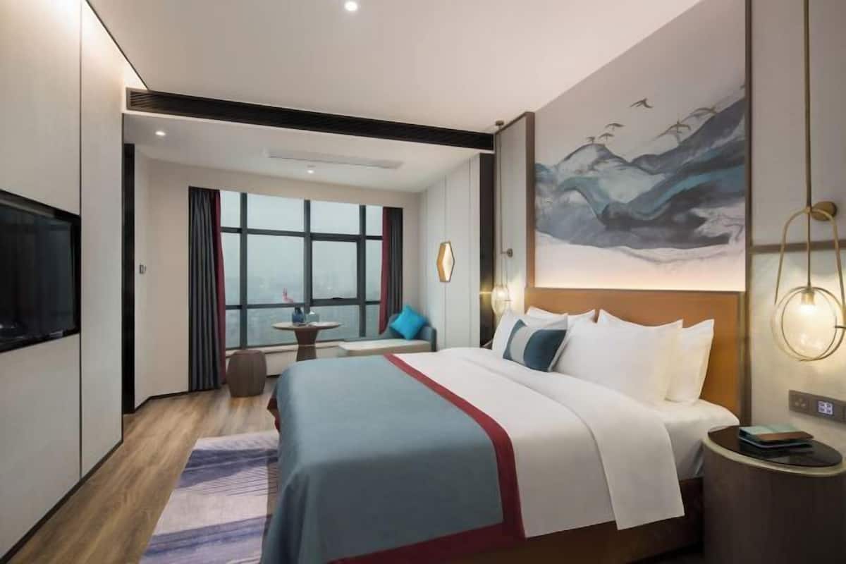 Ein modernes Hotelzimmer mit einem großen Bett, einem Fernseher, einem Gemälde an der Wand und einem Fenster mit Vorhängen.