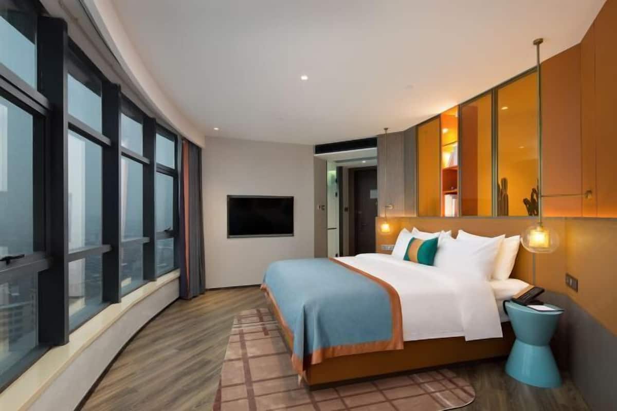 Ein modernes Hotelzimmer mit einem großen Bett, einem Fernseher und Blick auf die Stadt durch raumhohe Fenster.