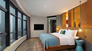 Quarto de hotel moderno com uma cama grande, televisão e vista para a cidade através de janelas do chão ao teto.