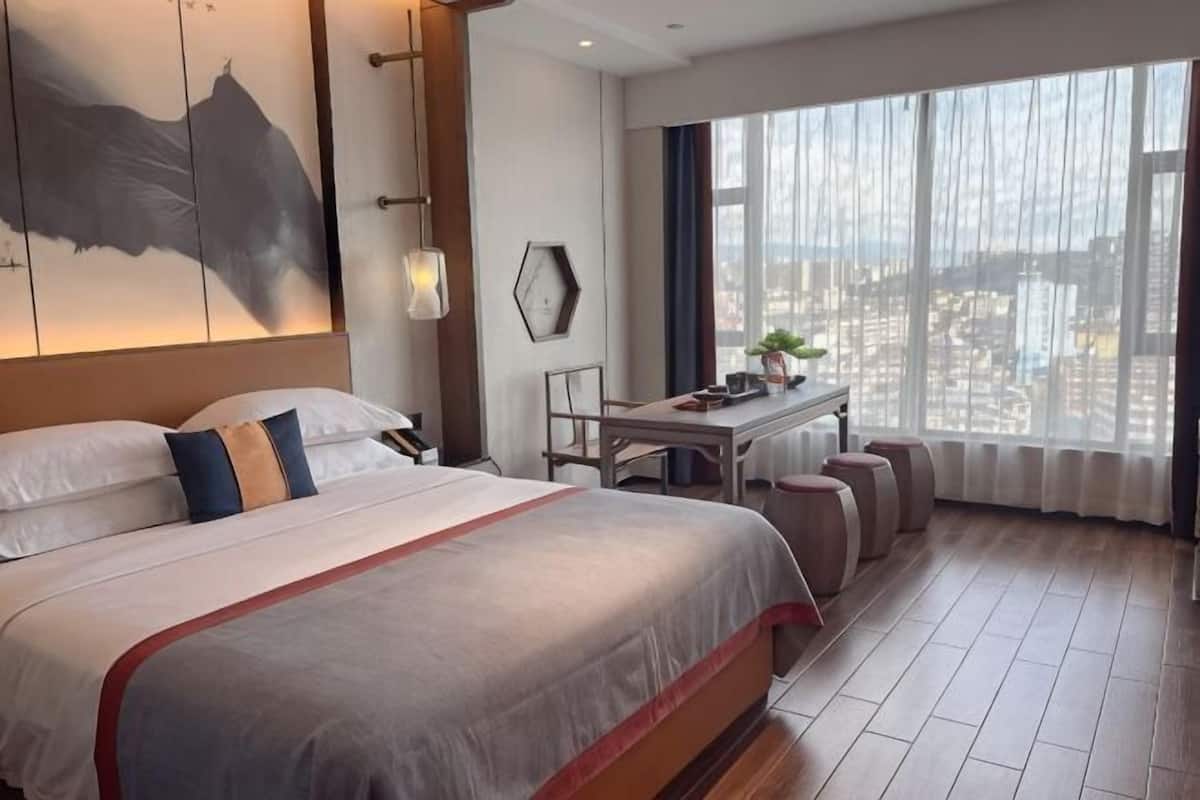 Ein Hotelzimmer mit einem großen Bett, einem Schreibtisch, einem Sessel und Blick auf die Stadt durch große Fenster.