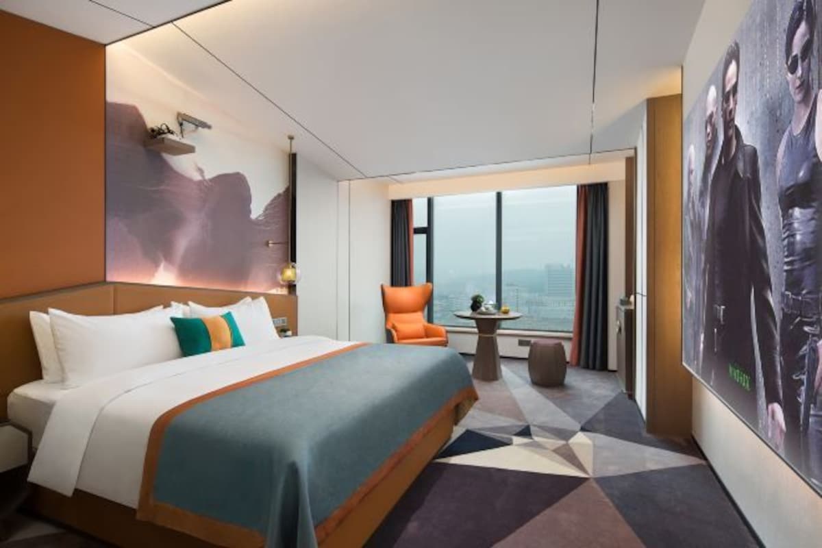 A modern hotel room with a large bed, a seating area with a chair and a small table, and a large window offering a city view.