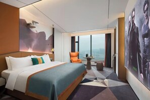 Une chambre d’hôtel dotée d’un grand lit, d’un coin salon avec un fauteuil et une petite table, et d’une grande fenêtre donnant sur la ville.