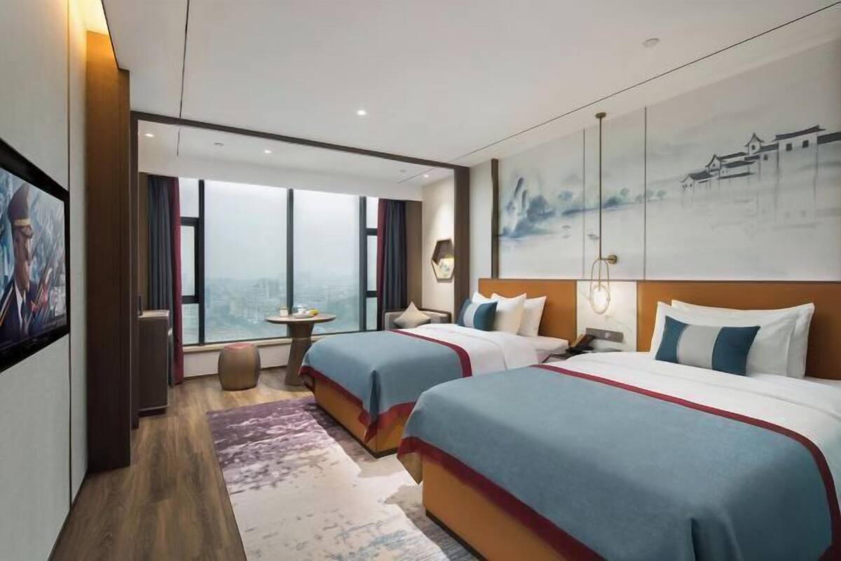 Ein Hotelzimmer mit zwei Betten, einem großen Fenster, einem Gemälde und Blick auf die Stadt.