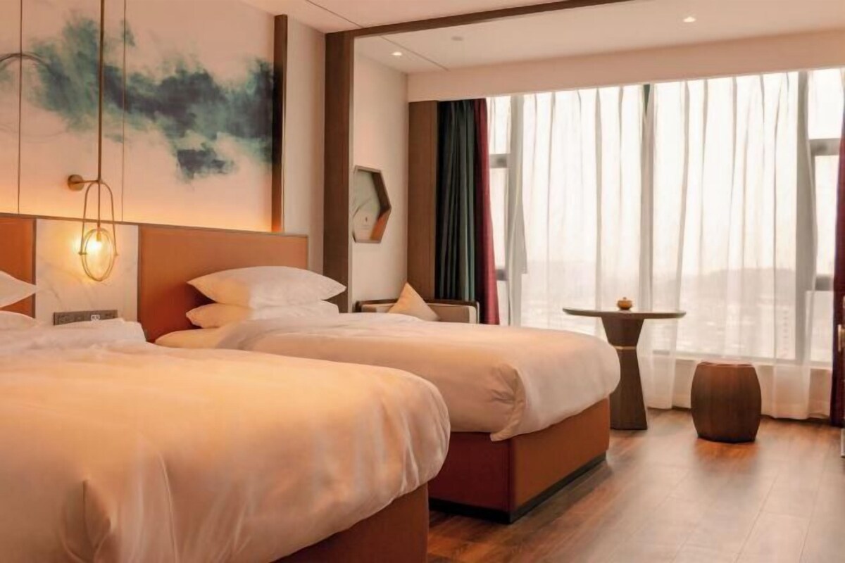 Une chambre d’hôtel avec deux lits, une petite table, une chaise et une grande fenêtre avec des rideaux.