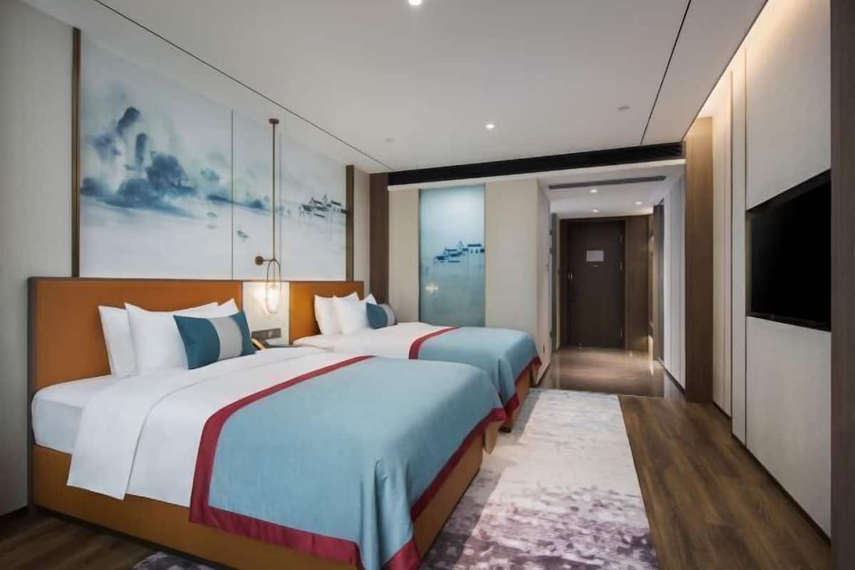 Ein Hotelzimmer mit zwei Betten, einem großen Gemälde an der Wand und einem an der Wand befestigten Fernseher.