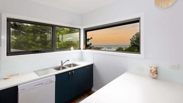 A modern kitchen with two large windows, a dishwasher, and a view of the ocean. - Jenkins Galwad-y-Mor | Beachfront Tomakin (TOMAKIN)
