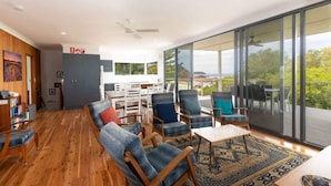 A modern living room with a wooden floor, a sofa set, a coffee table, and a view through large glass doors. - Jenkins Galwad-y-Mor | Beachfront Tomakin (TOMAKIN)