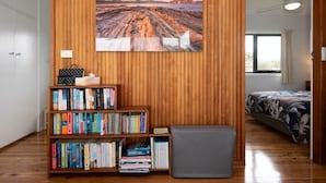 A room with a wooden wall, a bookshelf, a grey bench, and a bed with a floral bedspread. - Jenkins Galwad-y-Mor | Beachfront Tomakin (TOMAKIN)