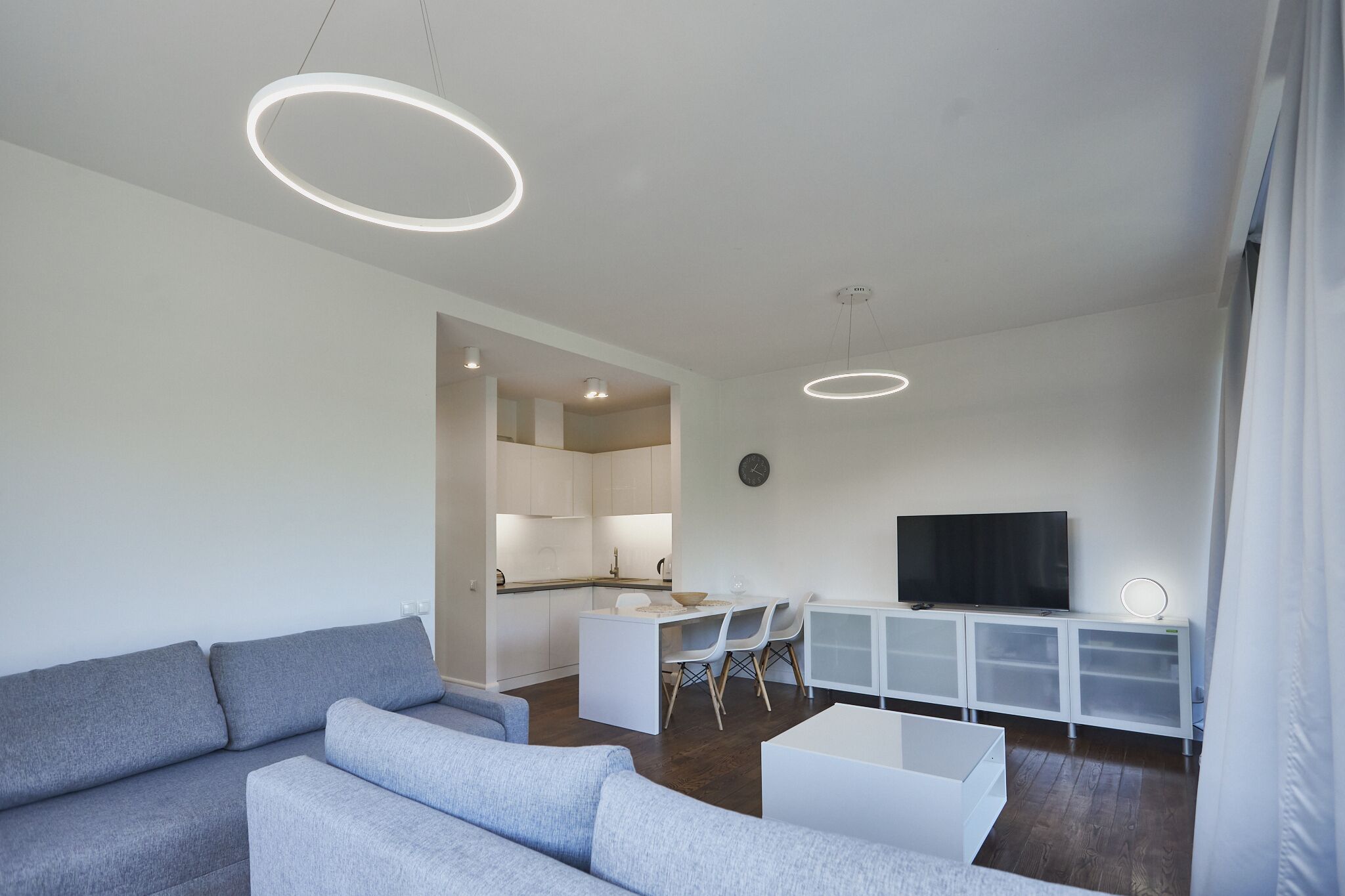 Una sala moderna con un sofá gris, un comedor con mesa y sillas, una cocina al fondo y un televisor sobre un mueble blanco.