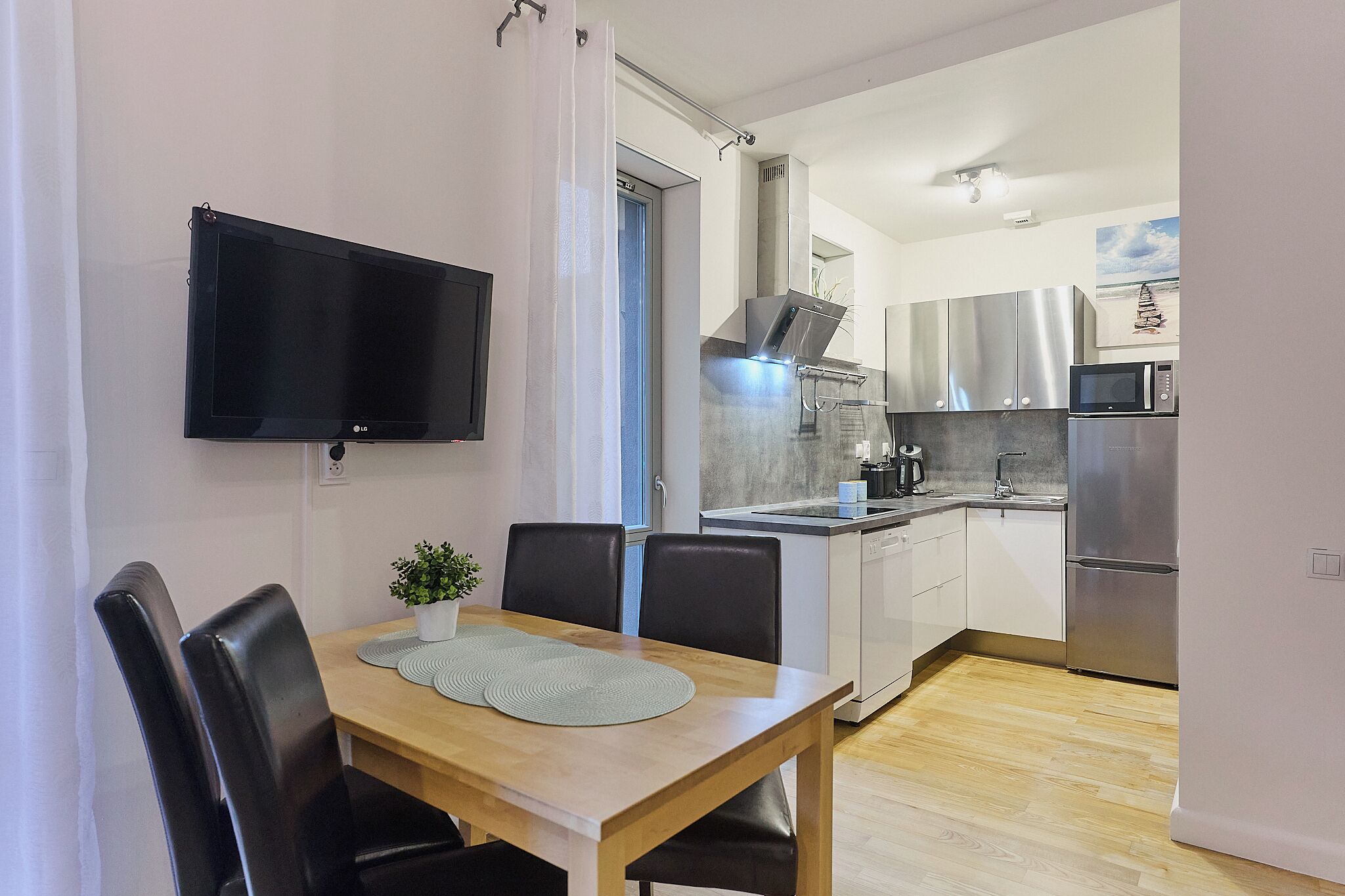 A modern kitchen with stainless steel appliances, a dining area with a wooden table and black chairs, and a flat-screen TV mounted on the wall.