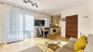 Una sala de estar moderna con un sofá, una mesa de centro y un televisor. Hay una zona de comedor con una mesa y sillas. El piso es de madera y hay un ventanal grande con cortinas.
