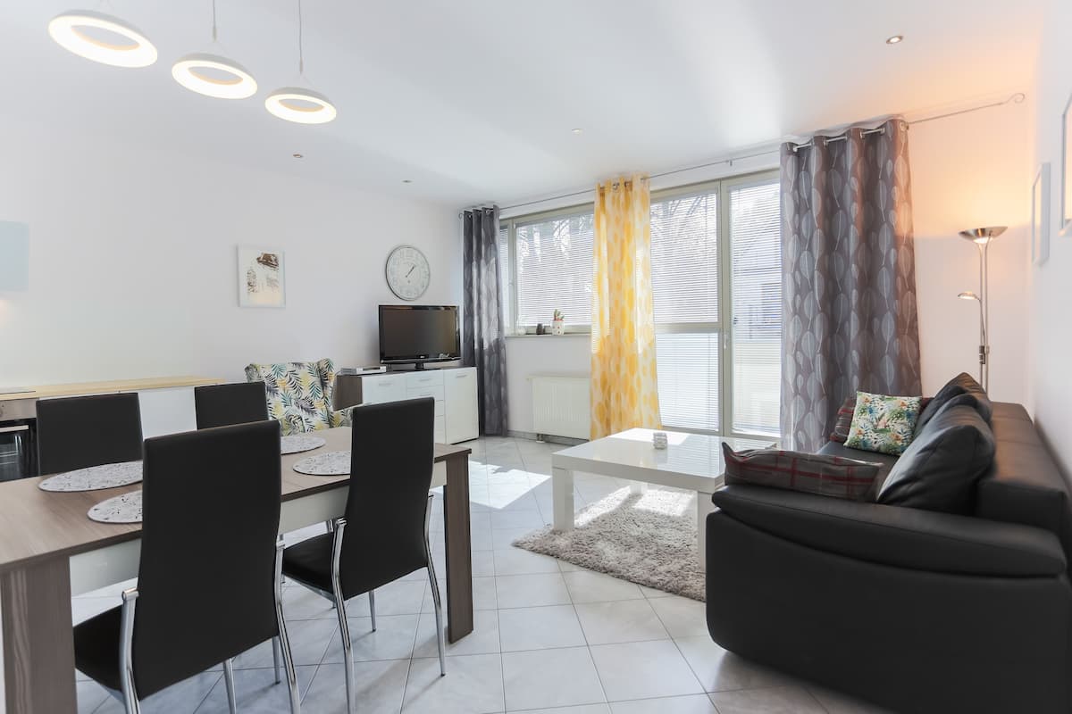 A modern living room with a sofa, dining table, and chairs. There is a TV on a stand, a clock on the wall, and a window with curtains.