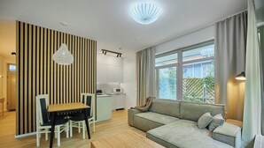 A modern living room with a sofa, dining table, and chairs, a kitchen area, and large windows with curtains. - Apartamenty Swinoujscie - Marinos (Swinoujscie)