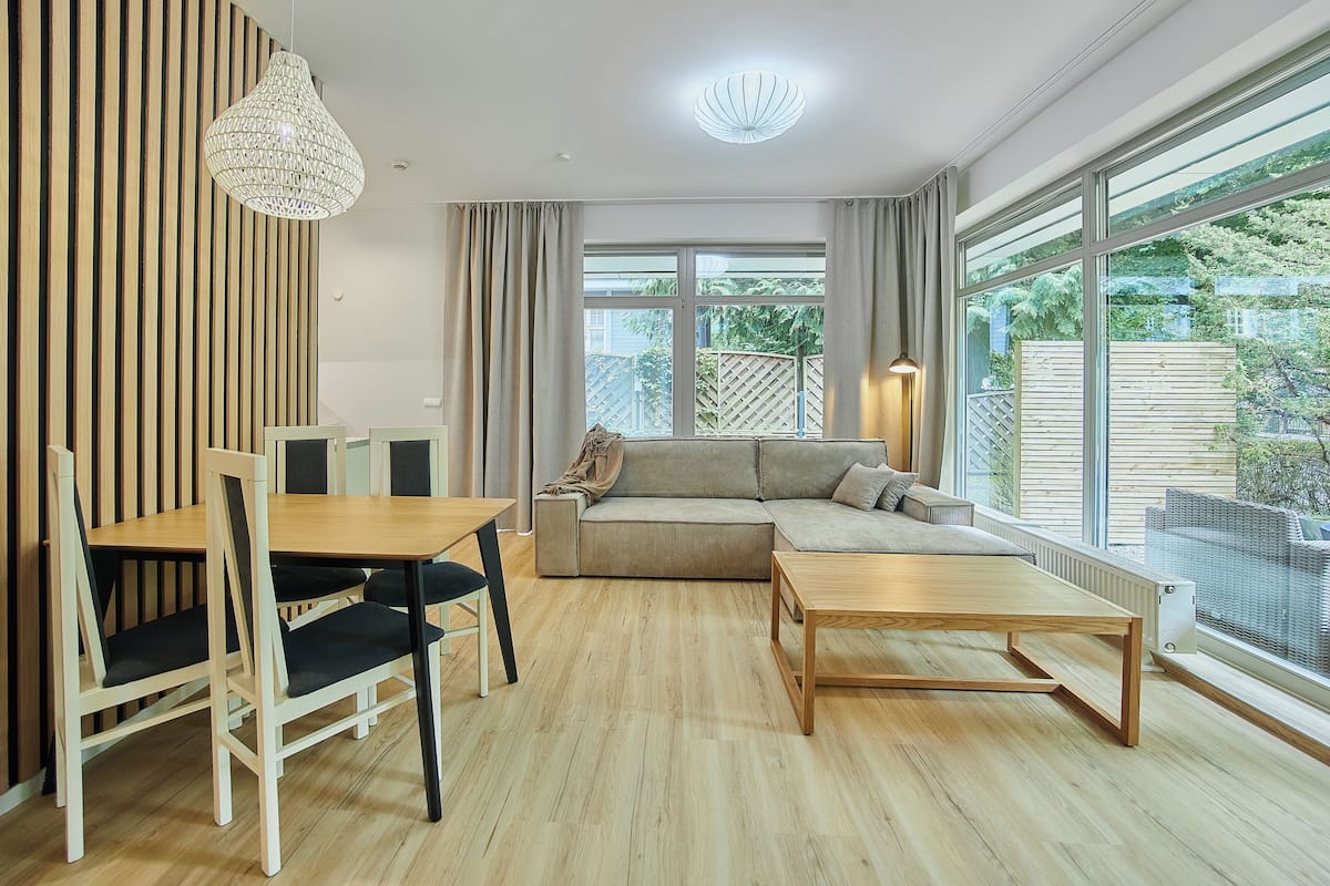 A modern living room with a wooden floor, a sofa, a dining table with chairs, and a large window with a view of greenery.